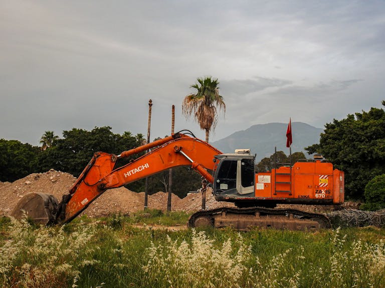 A bright orange excavator at a construction site surrounded by greenery in Payas, Hatay, Türkiye.